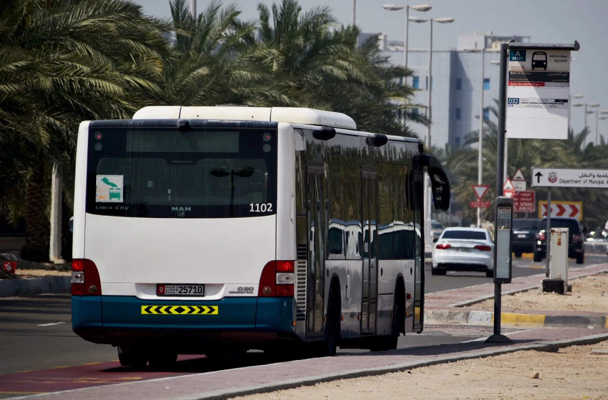 A white and teal city bus beside a bus stop on a palm-lined Abu Dhabi road