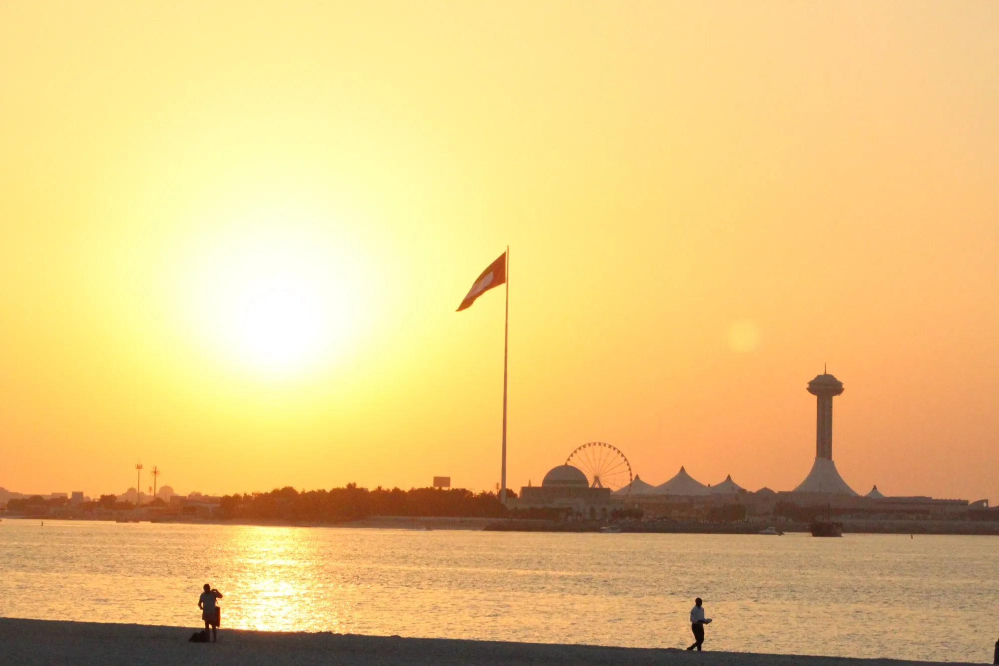 Sunset over the water from Abu Dhabi Corniche beach, with a tall flagpole and the Marina Mall tower across the bay