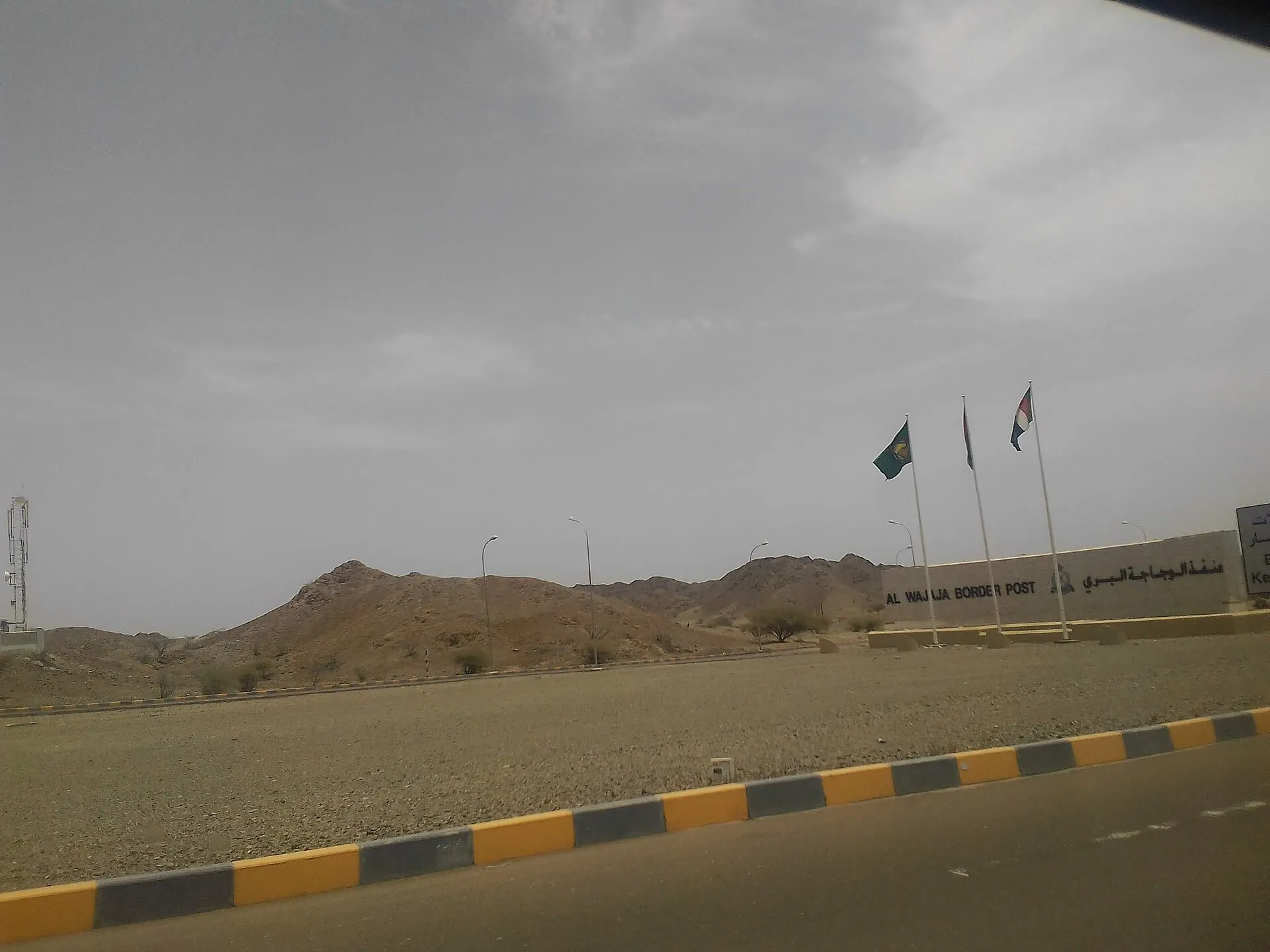 The Al Wajajah border post sign in Oman, with three flags and rocky hills behind, seen from a passing car