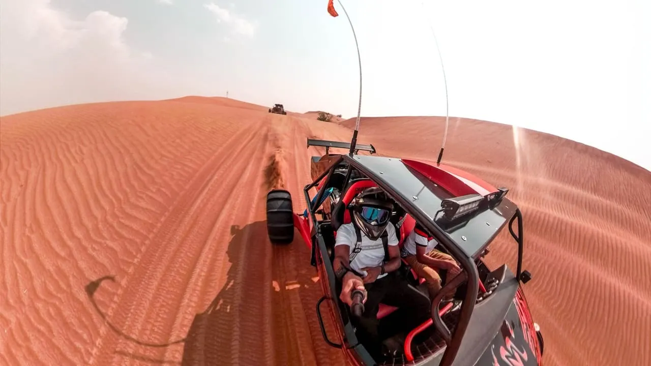 A buggy driver in a helmet and goggles following another buggy across red dunes in the Dubai desert