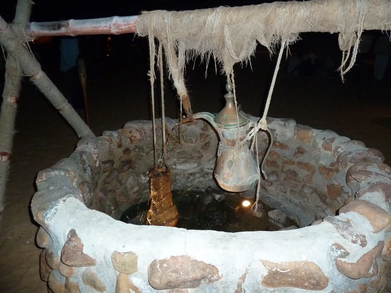 A traditional stone well with a brass coffee pot hanging above it at a Dubai desert camp at night