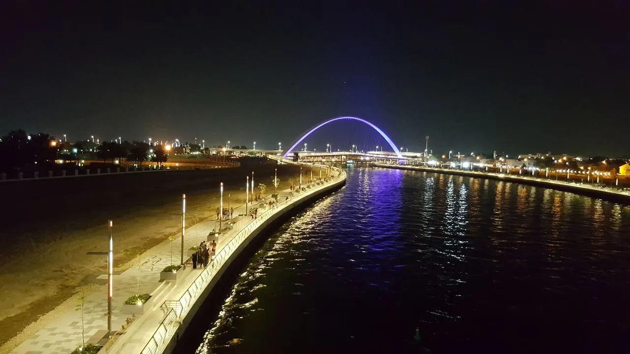 The Dubai Water Canal and its waterside walkway at night, with a lit arch footbridge in the distance