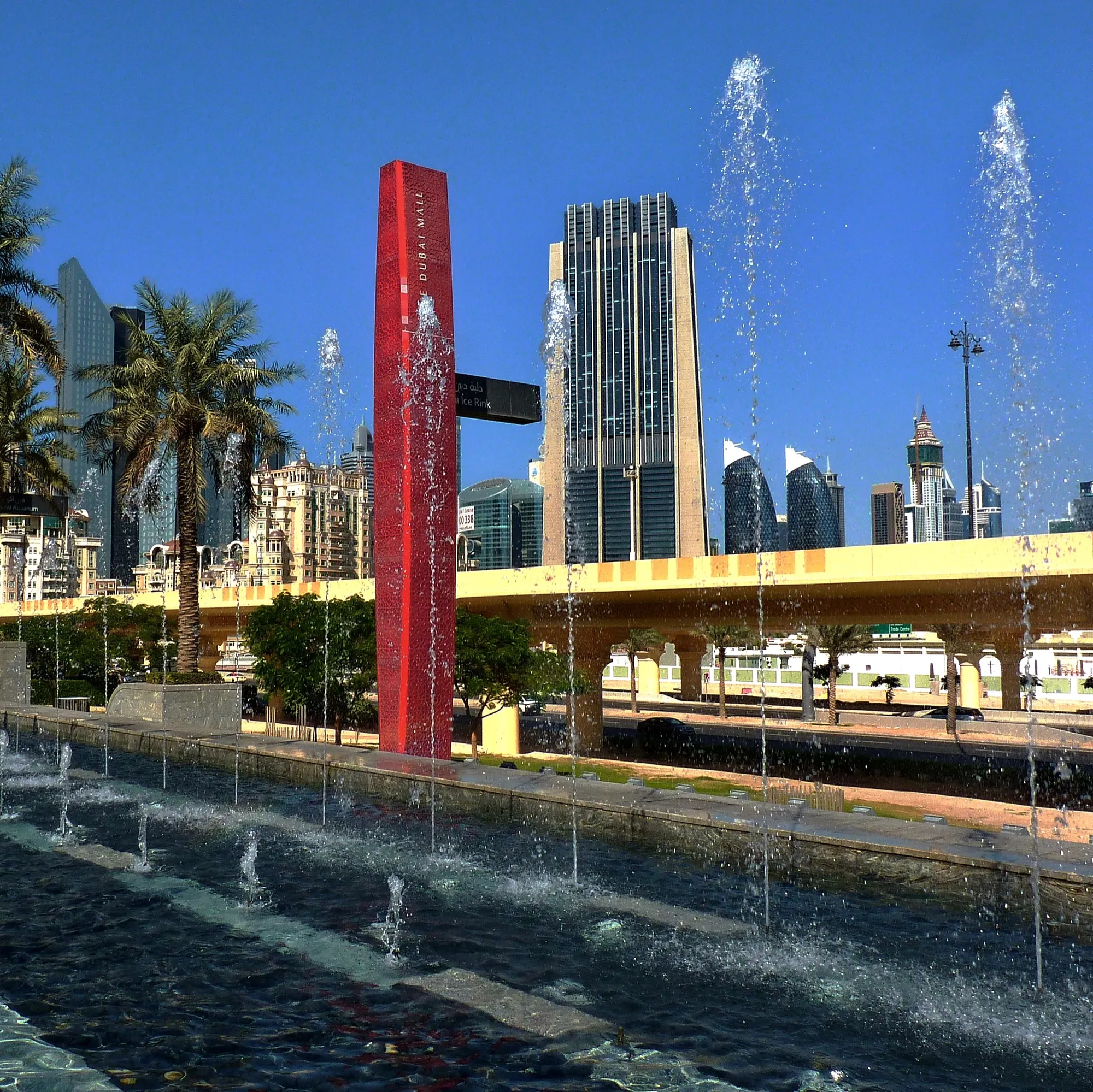 Water jets and a red Dubai Mall marker tower beside a raised road outside the mall