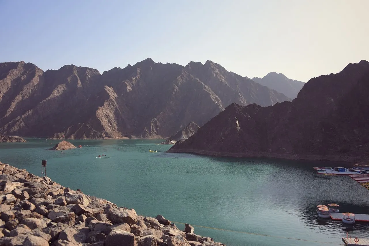 The green water of Hatta Dam between the bare peaks of the Hajar Mountains, with pedal boats moored on the right