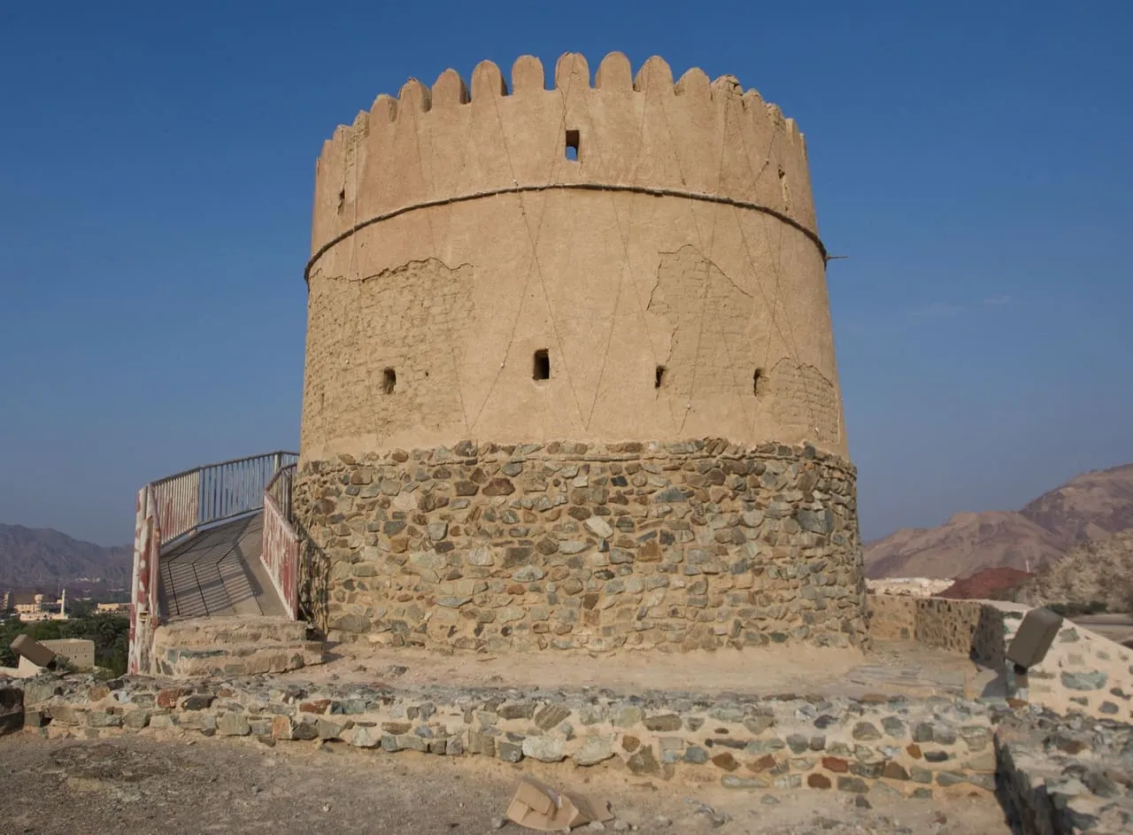A round stone and mud watchtower on a hilltop in Hatta, with mountains behind