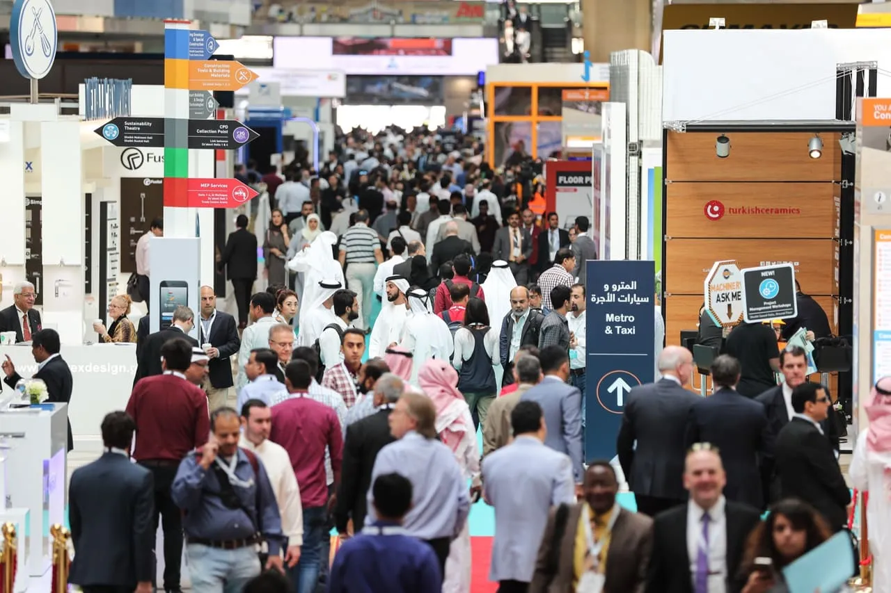 Crowded hall at a large Dubai trade exhibition with directional signs and a metro and taxi sign