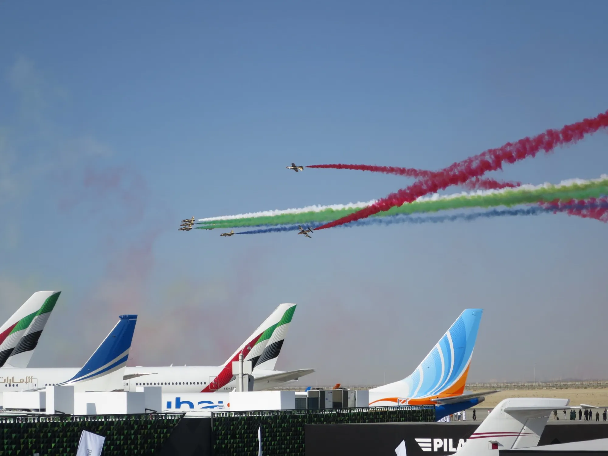 The Al Fursan display team trailing red, green, white and black smoke over aircraft tails at the Dubai Airshow 2023