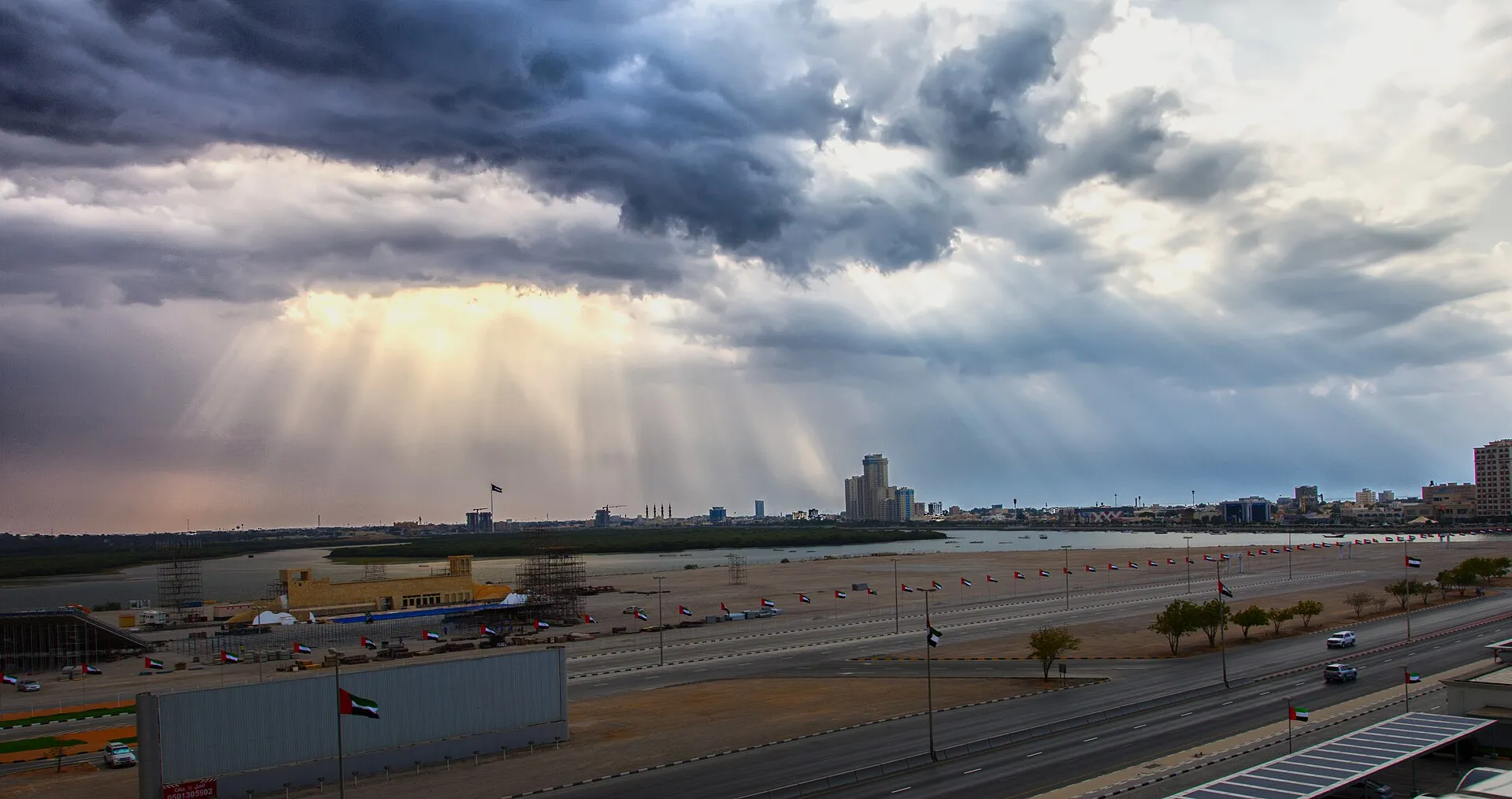 Ras Al Khaimah city seen across the creek under a stormy sky, with UAE flags lining the road in the foreground