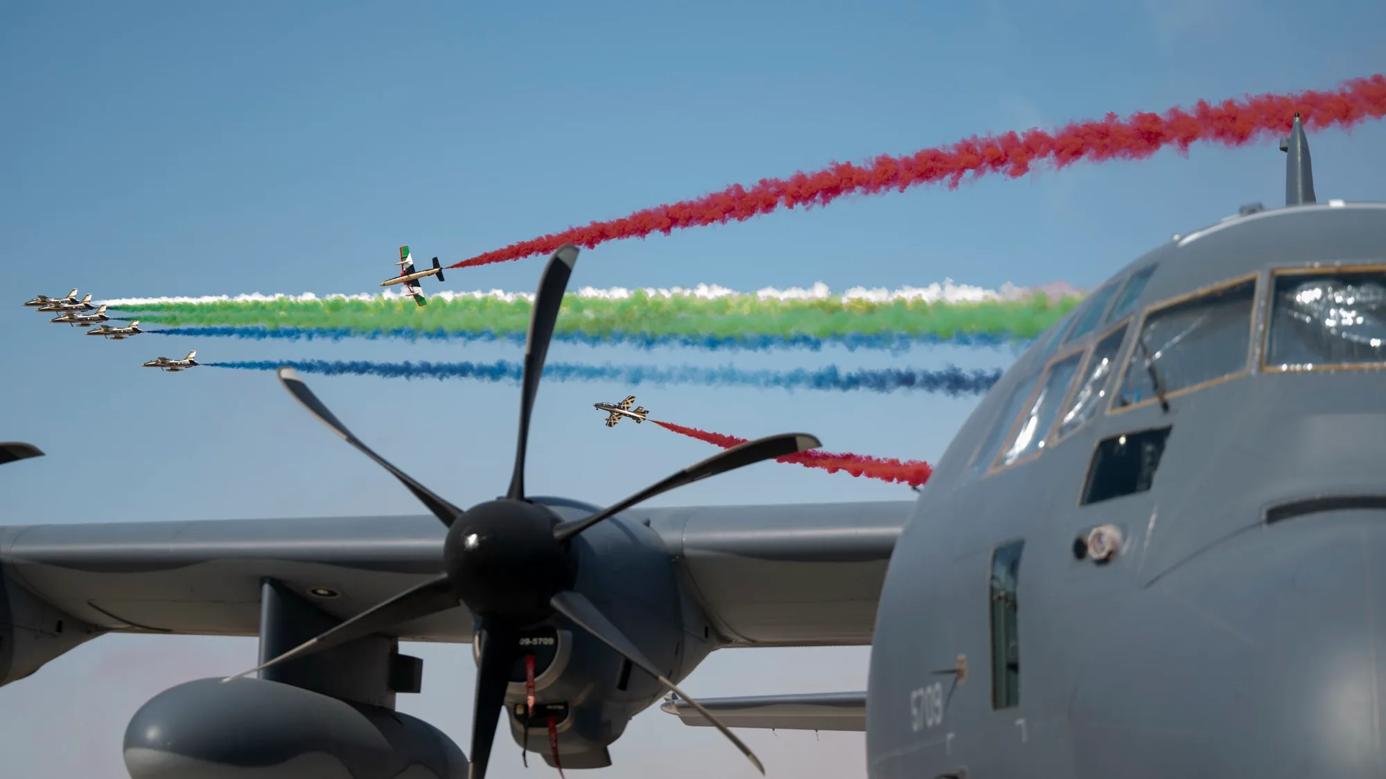 The UAE display team Al Fursan trailing red, green, white and blue smoke over a parked HC-130J at the 2023 Dubai Airshow, Al Maktoum International Airport