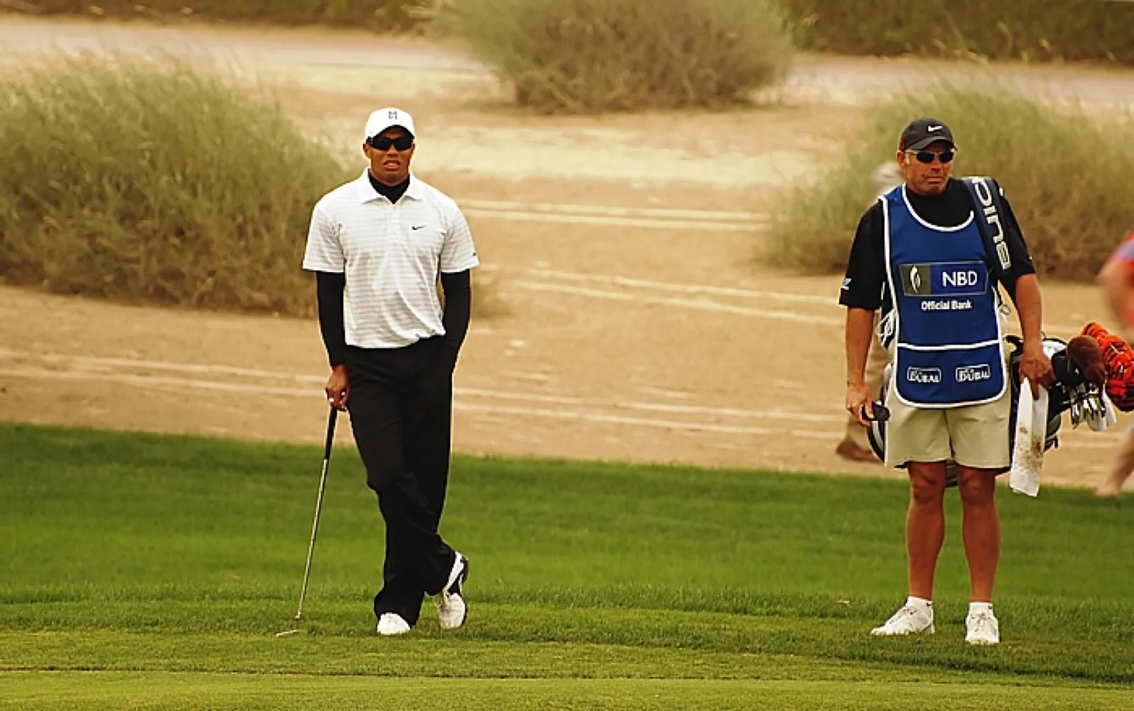 A golfer and his caddie waiting on the fairway during the Dubai Desert Classic, with desert scrub behind