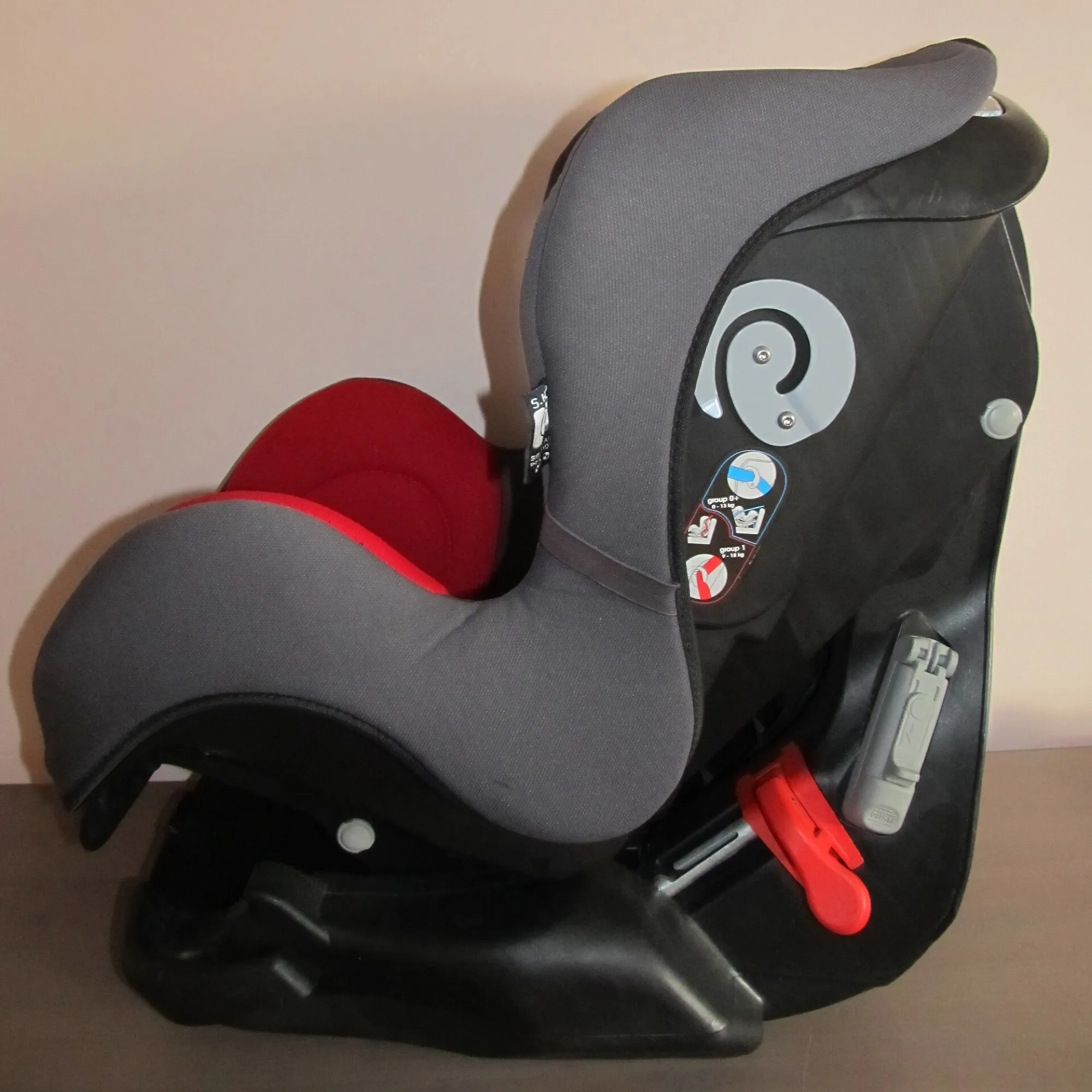 A convertible child car seat in grey and red, seen from the side, with its belt guides and base
