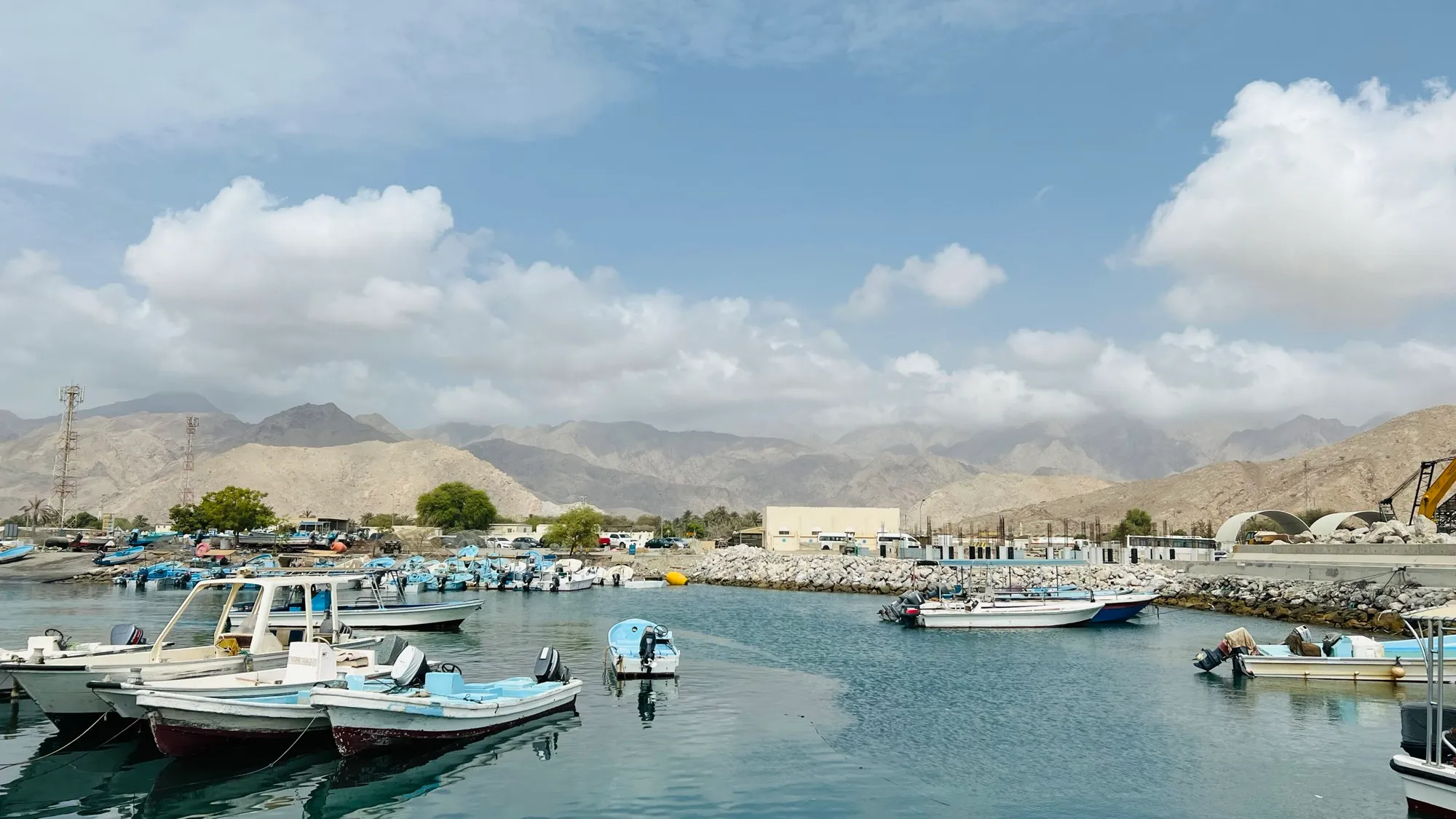 Small fishing boats moored in the harbour at Dibba on the Fujairah coast, with the Hajar Mountains rising behind under broken cloud