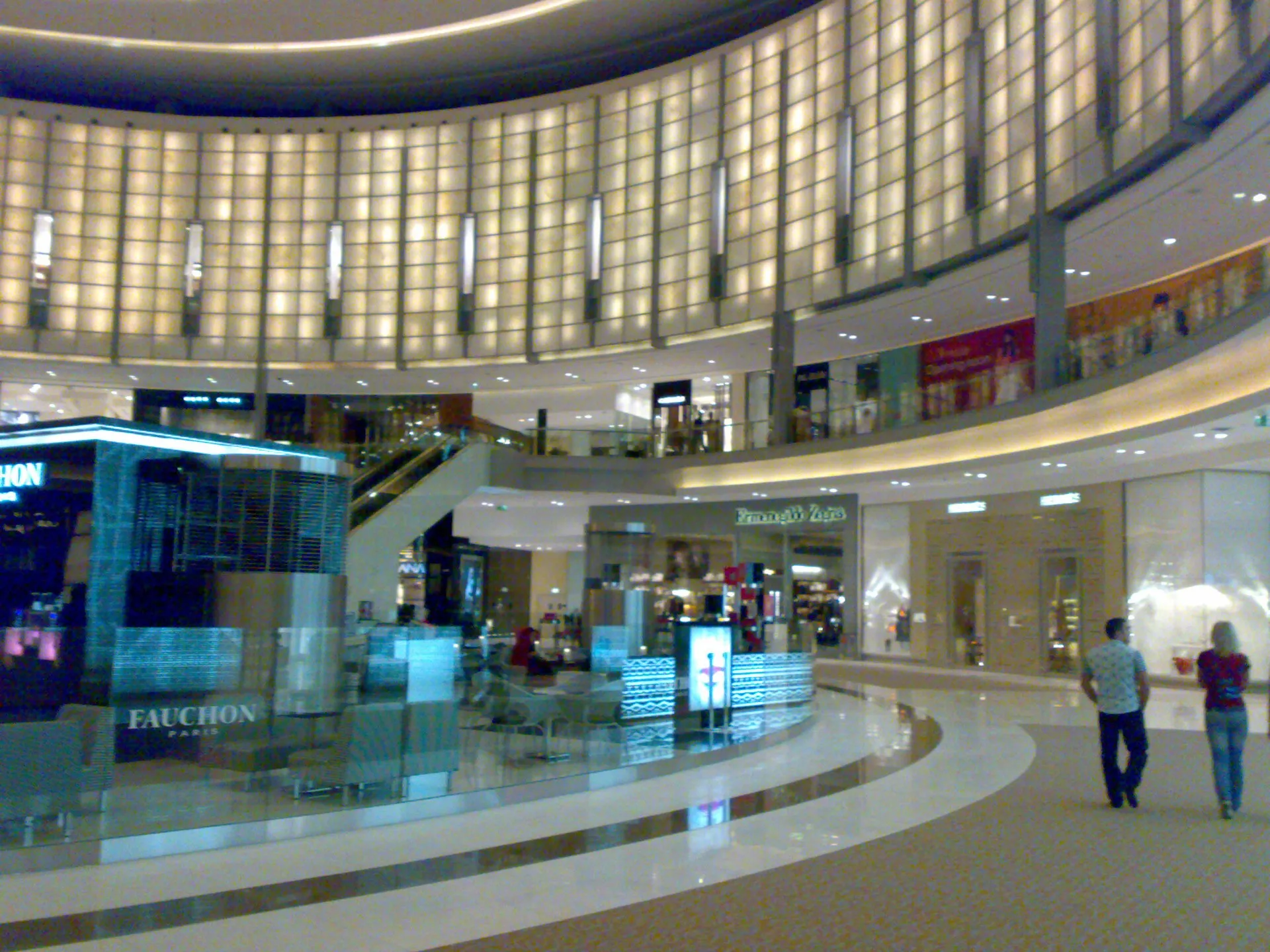 A curved, lit atrium inside The Dubai Mall with luxury store fronts and a cafe on the ground floor