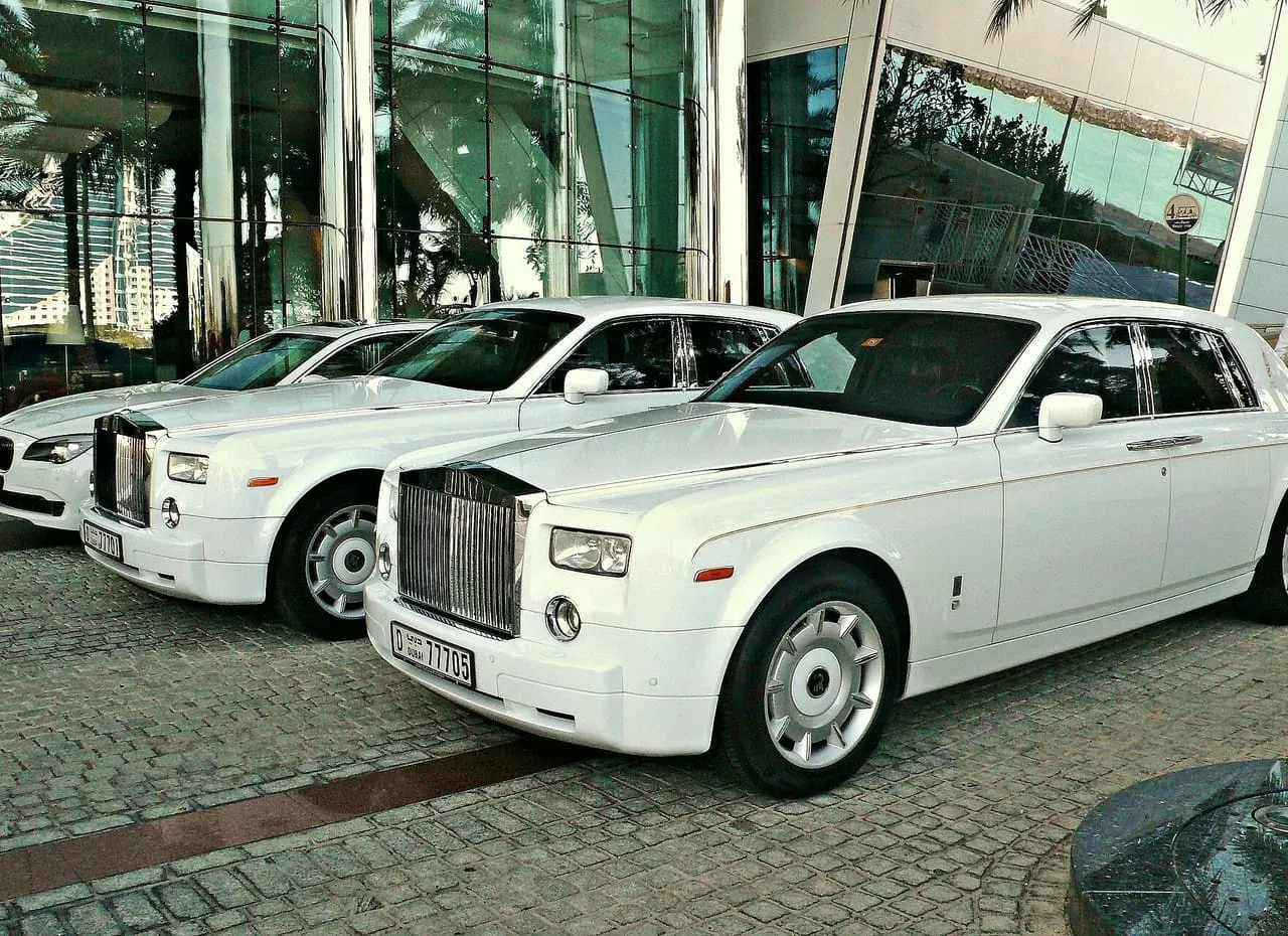 Two white Rolls-Royce Phantoms and a white BMW sedan with Dubai number plates, parked outside a glass-fronted building in Dubai