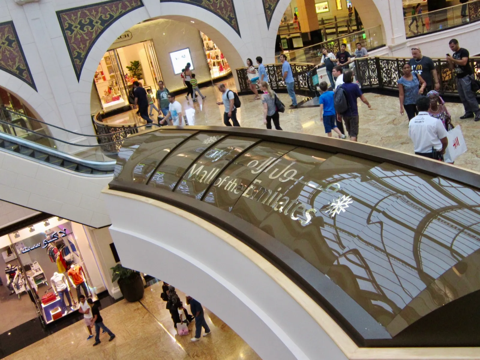 Shoppers on the marble floors of Mall of the Emirates, seen from an upper level past a curved sign