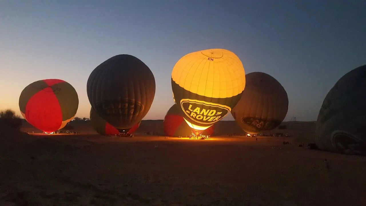 Hot air balloons being inflated on the desert floor outside Dubai before sunrise, one glowing from its burner