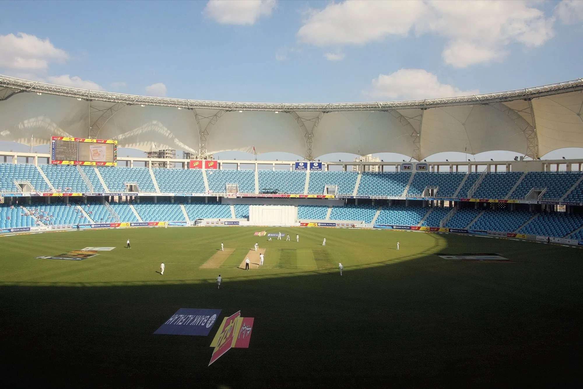 Inside Dubai International Cricket Stadium in Dubai Sports City during a Test match, with blue seating and the white tensile roof