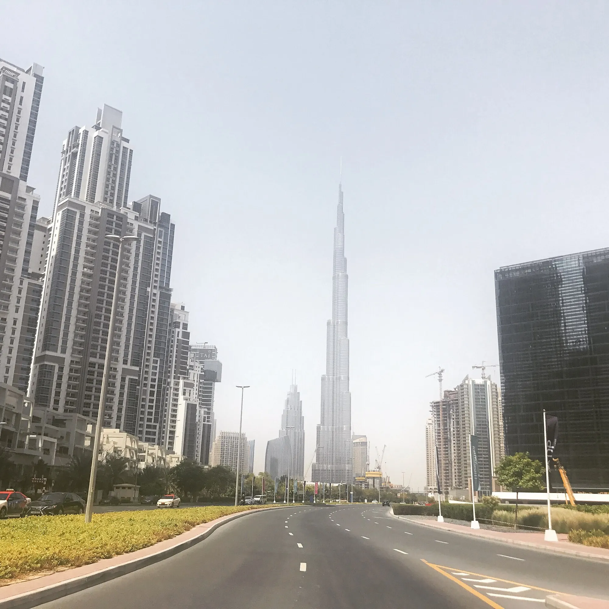 A wide road in Business Bay, Dubai, with the Burj Khalifa rising between office and residential towers