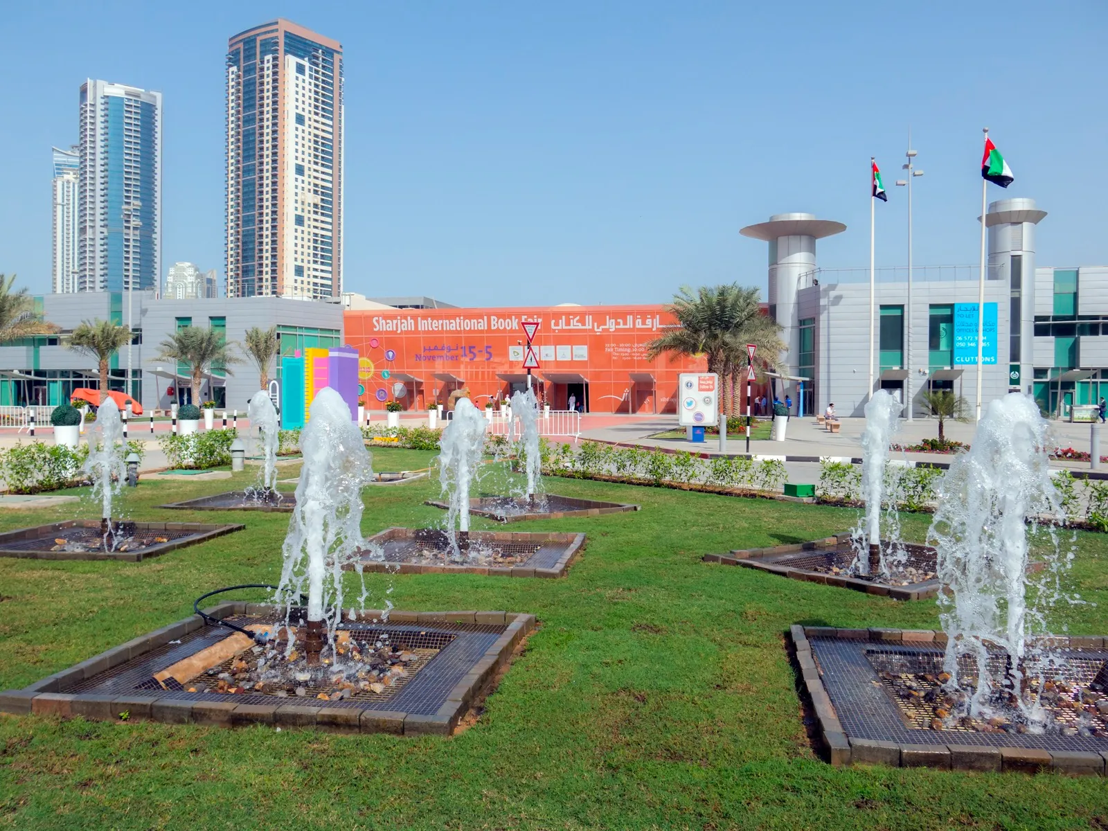 The Sharjah International Book Fair entrance hall at Expo Centre Sharjah, with fountains on the lawn and UAE flags