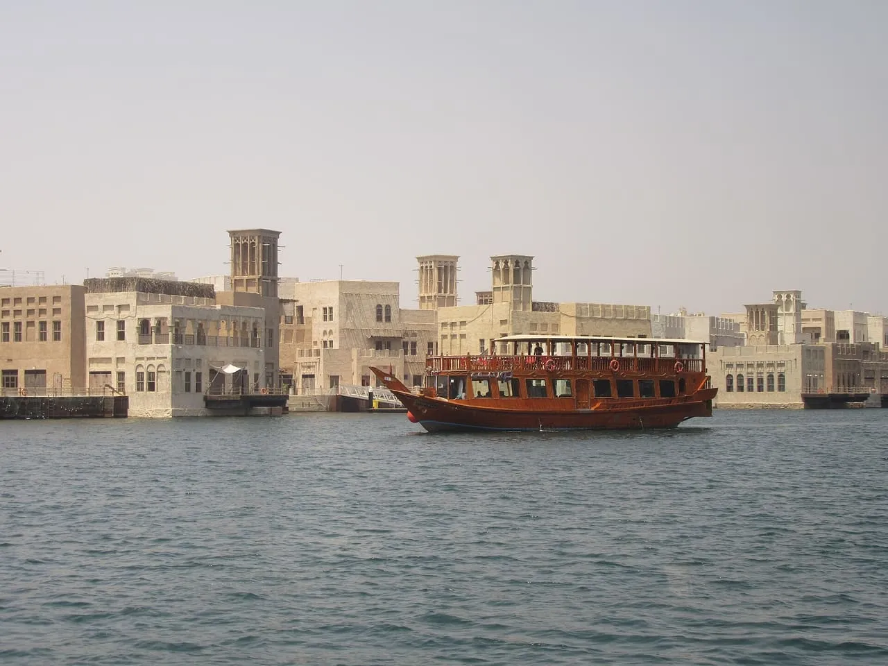 A two-deck wooden cruise dhow on Dubai Creek, passing wind-tower buildings on the Bur Dubai bank