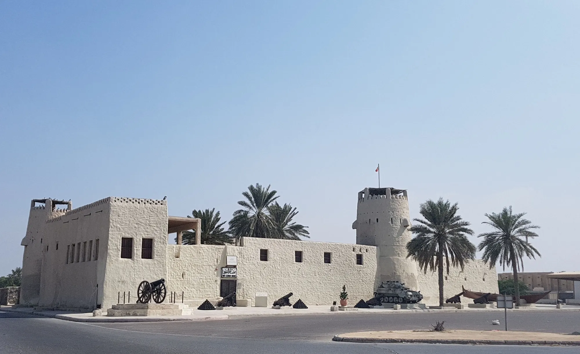 Whitewashed Umm Al Quwain Fort with cannons on display, a round corner tower and date palms under a clear sky