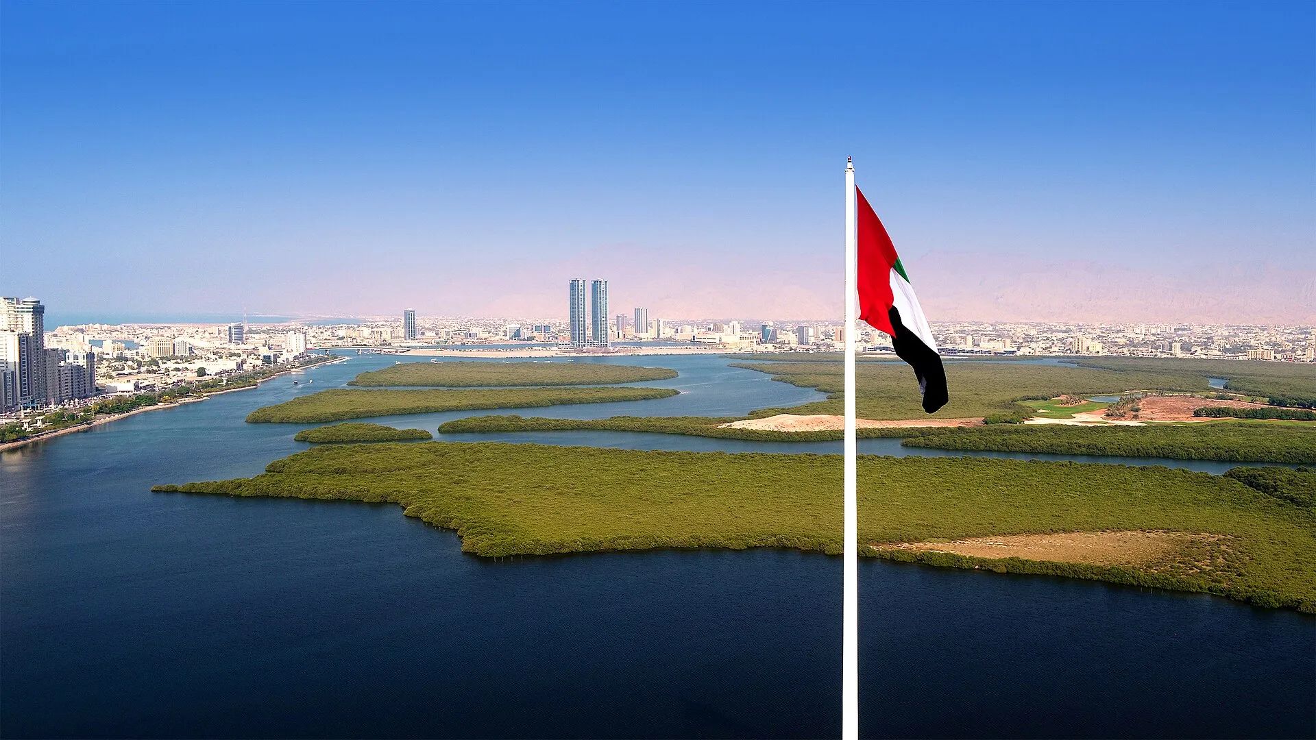 Aerial view of Ras Al Khaimah city and its mangrove creek, with a UAE flag on a tall pole in the foreground