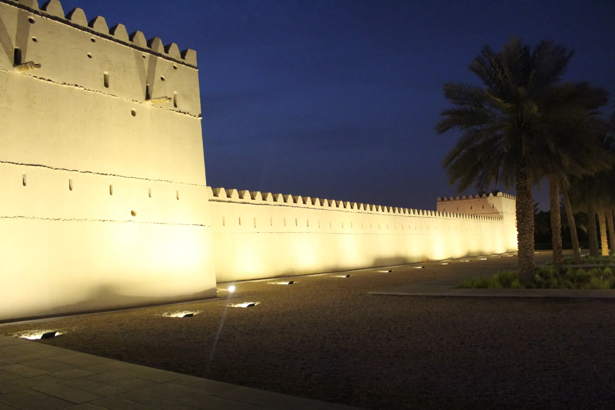 The floodlit mud-brick walls and corner tower of Qasr Al Muwaiji in Al Ain at dusk, with date palms to the right