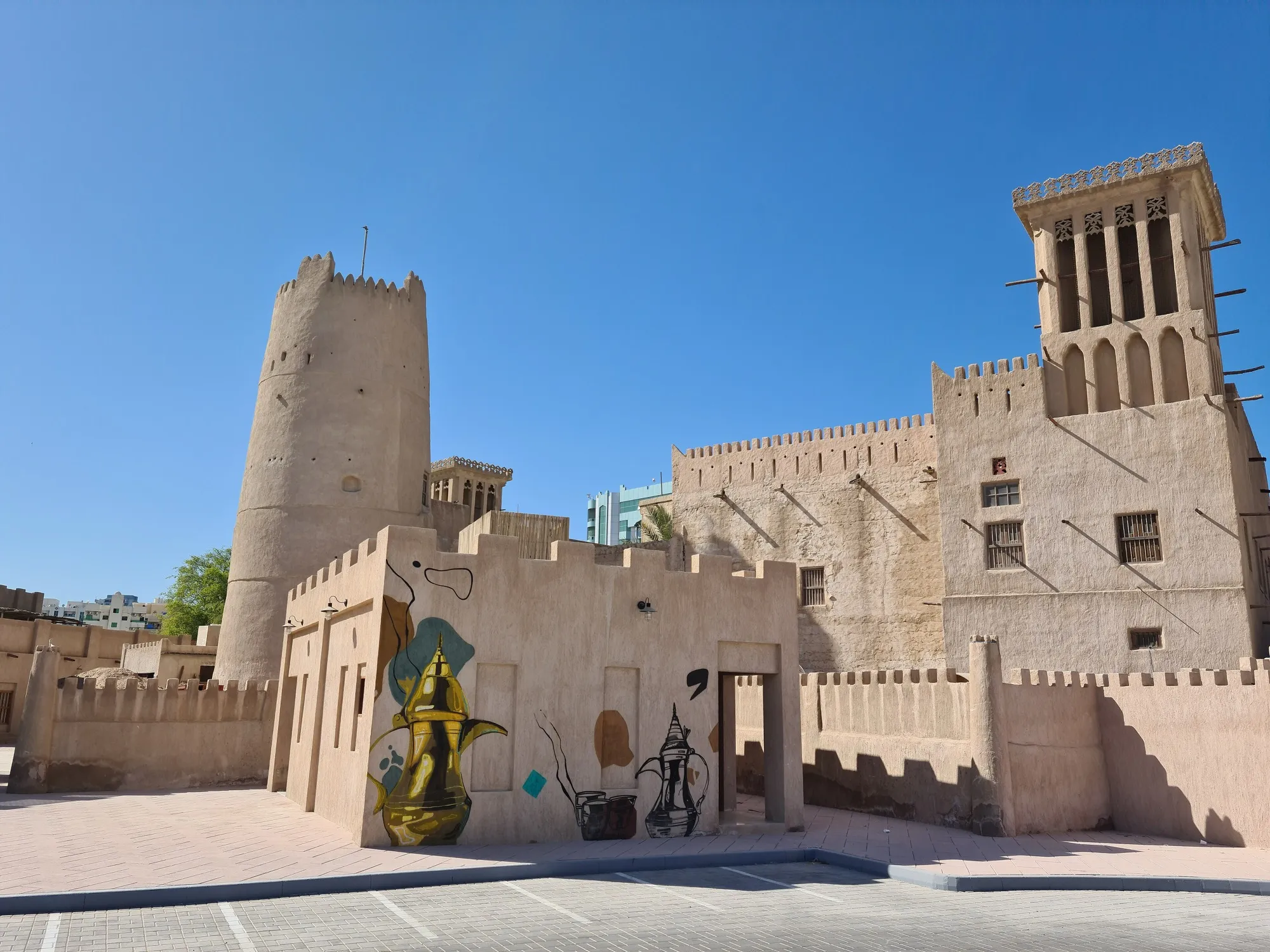 The round watchtower and a wind tower of the old fort in Ajman, with a coffee-pot mural in the foreground
