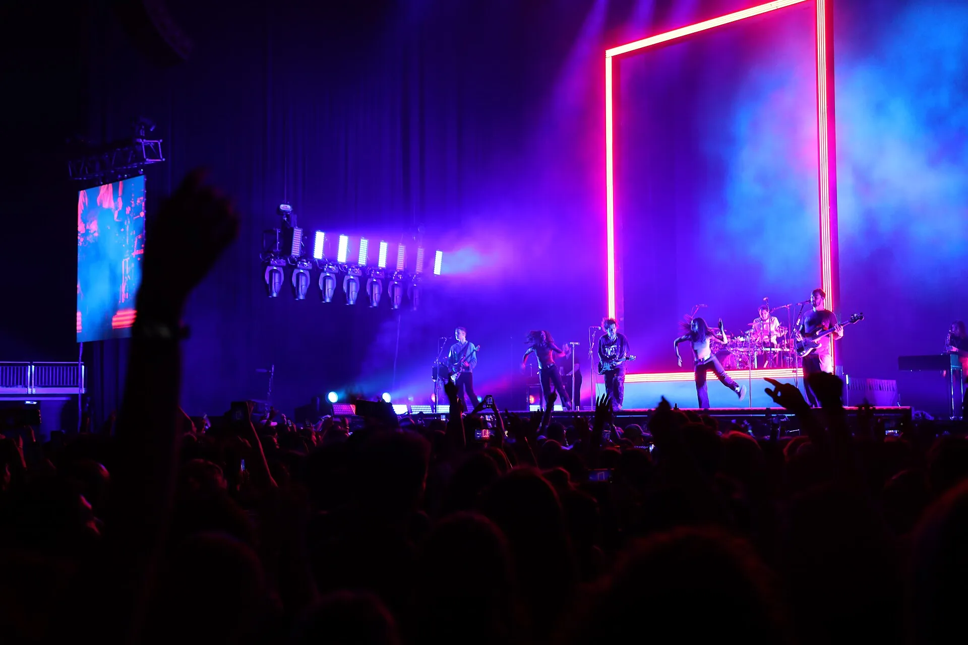 Band performing on a lit stage in front of a large neon frame, with a crowd in the foreground