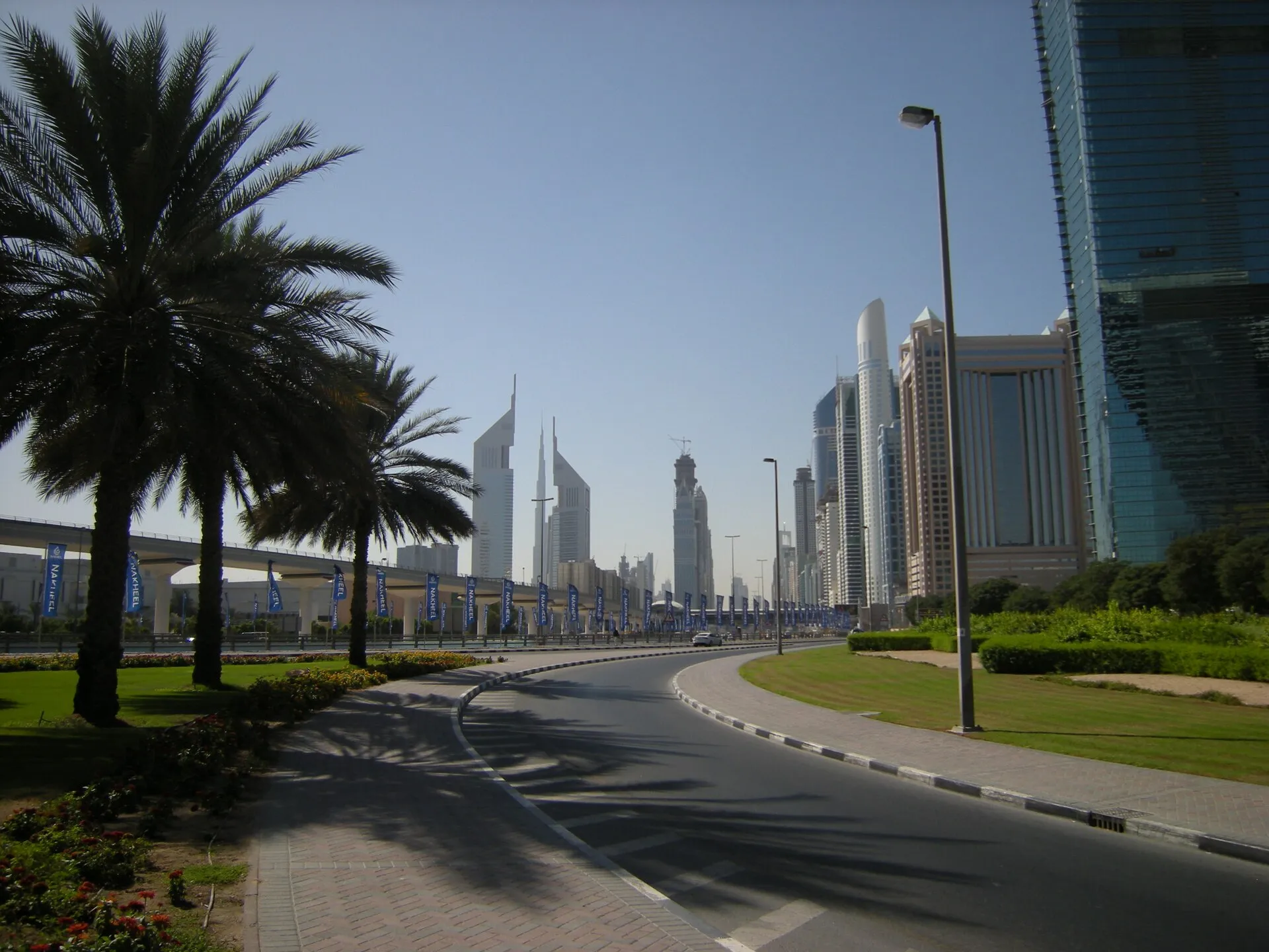 A curved road lined with palm trees beside the Dubai Metro viaduct, with the Emirates Towers and other Sheikh Zayed Road high-rises in the distance