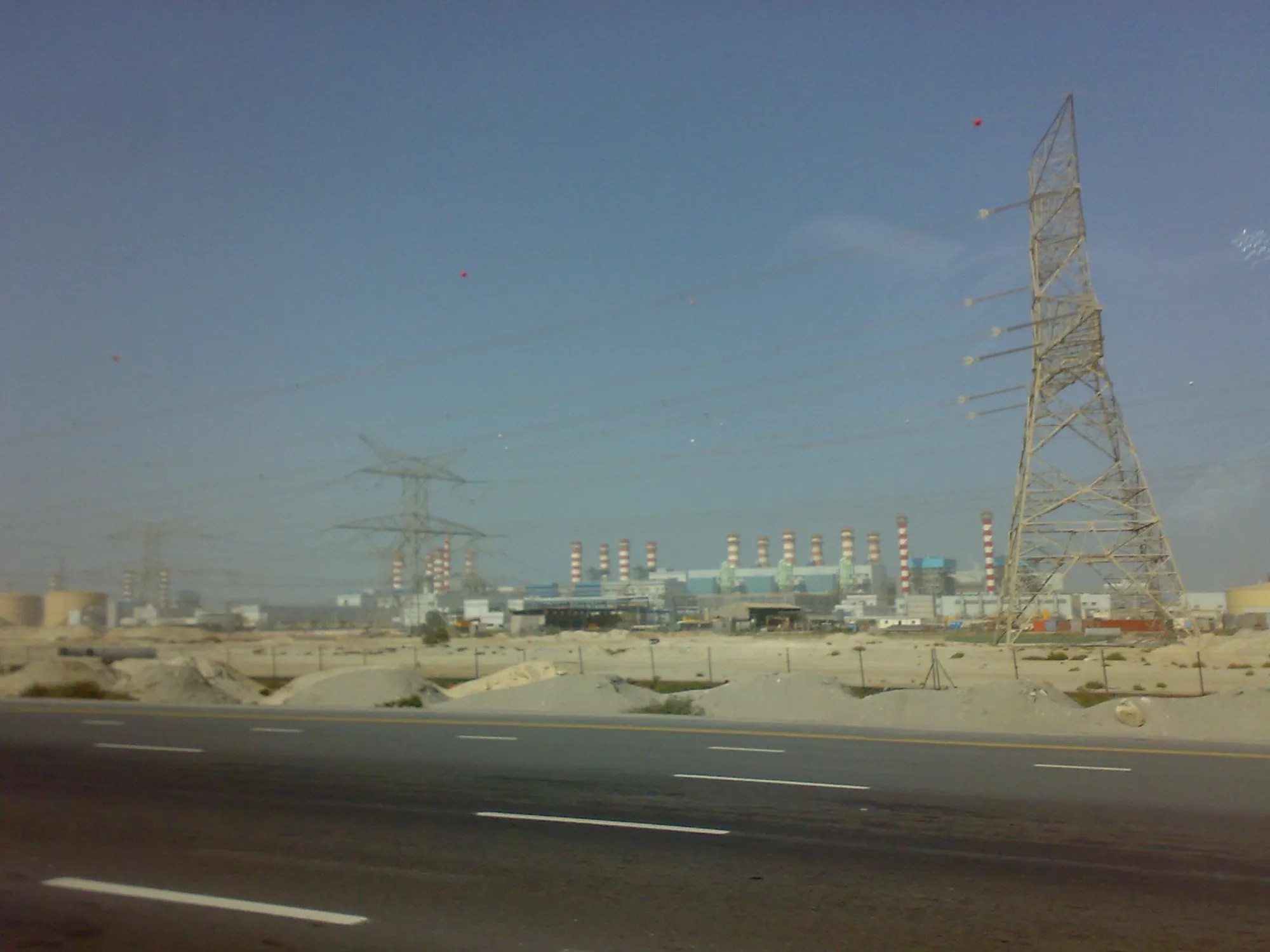 A multi-lane road beside open sand in the Jebel Ali area, with the stacks of a power station and pylons behind