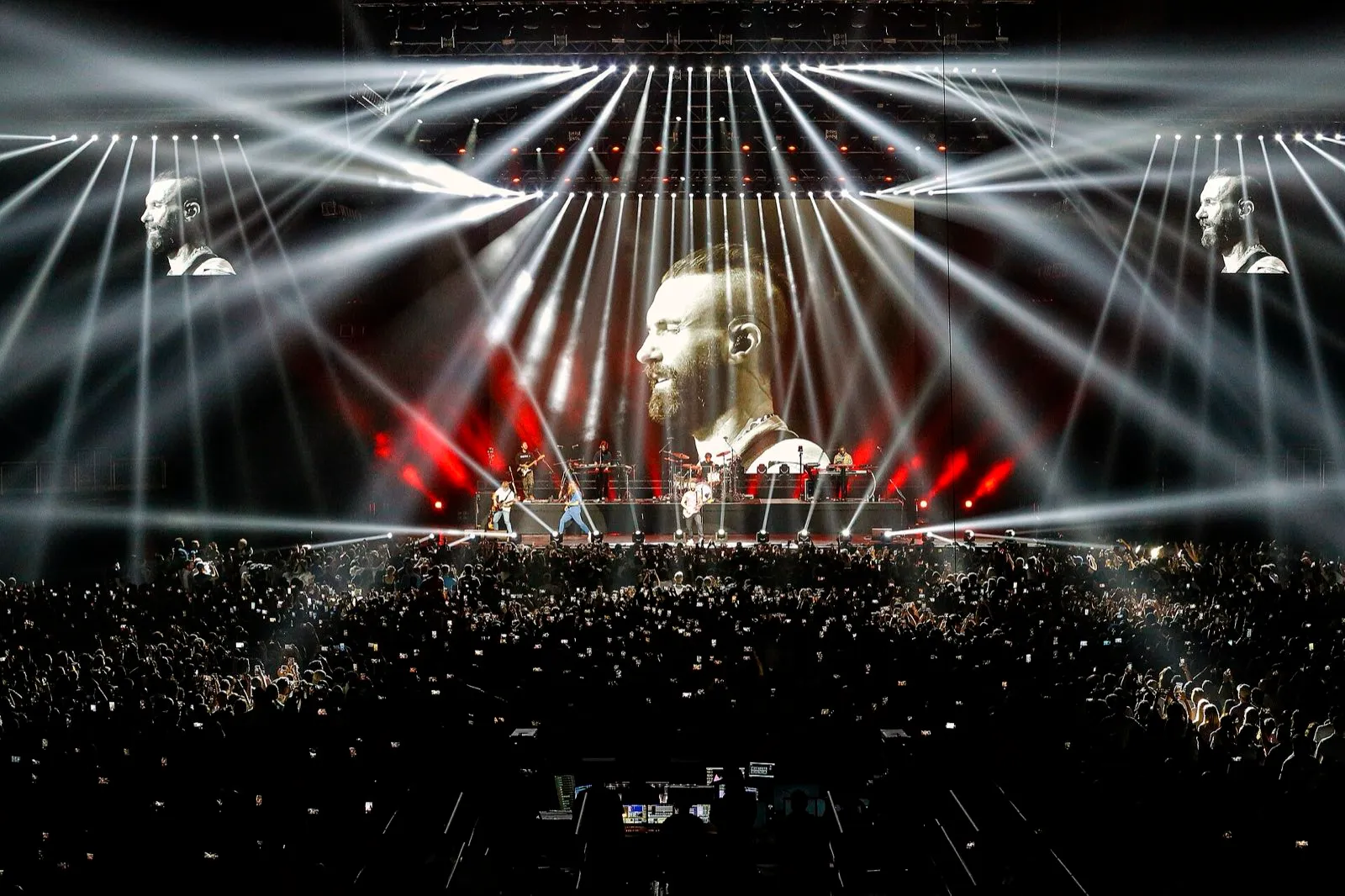 A band on a wide arena stage with a large video screen and beams of white light above a packed crowd