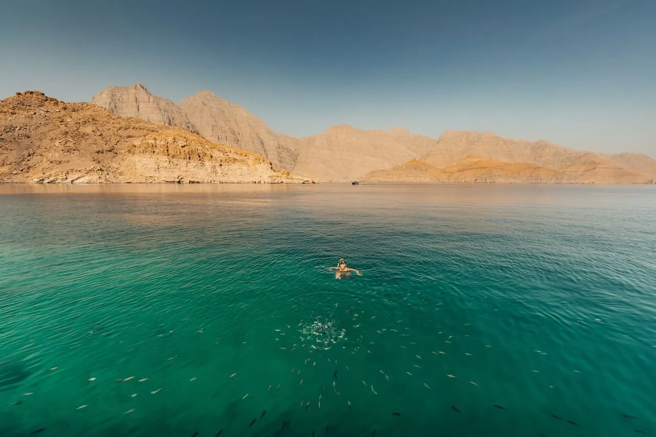 A swimmer in clear green water at a stop in Khor Ash Sham, with fish below and the Musandam mountains behind