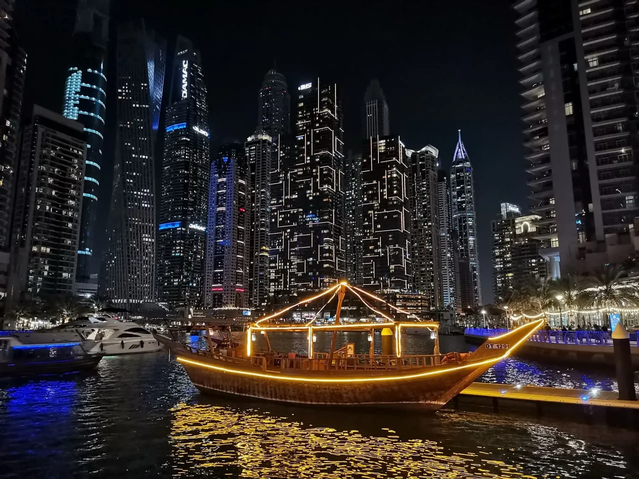 A wooden dinner dhow lit with lights at a pontoon in Dubai Marina at night, with the marina towers behind