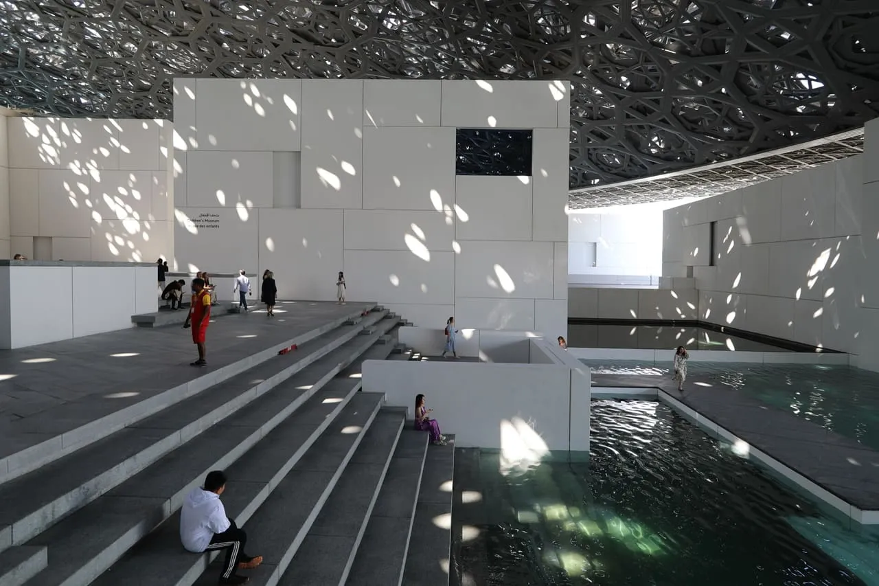 Steps, water and white gallery blocks under the latticed dome of Louvre Abu Dhabi on Saadiyat Island