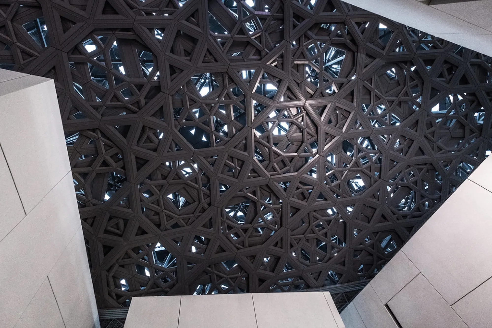 Looking up at the layered geometric lattice of the Louvre Abu Dhabi dome, with light coming through the gaps