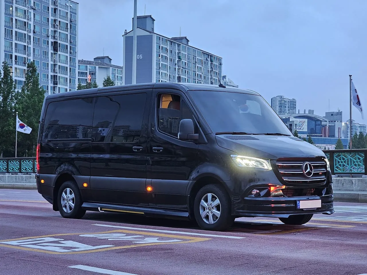 A black Mercedes-Benz Sprinter Tourer passenger van with tinted windows, on a city road at dusk