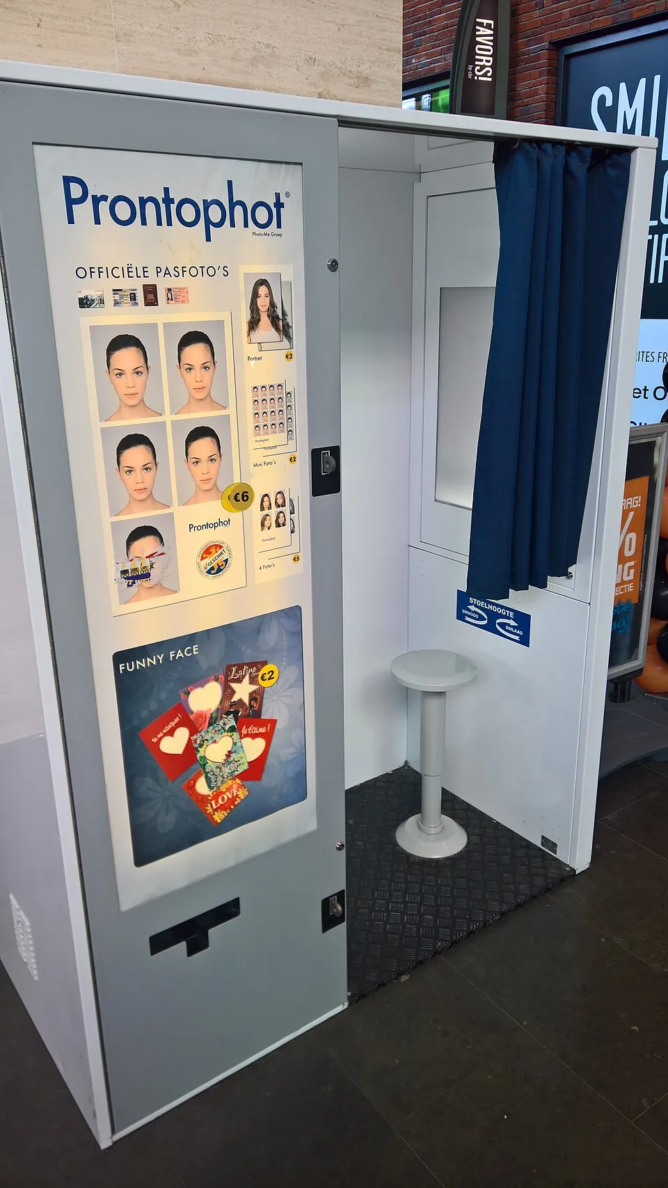 A passport photo booth with a white interior and a stool, beside a sample sheet of passport-style photos