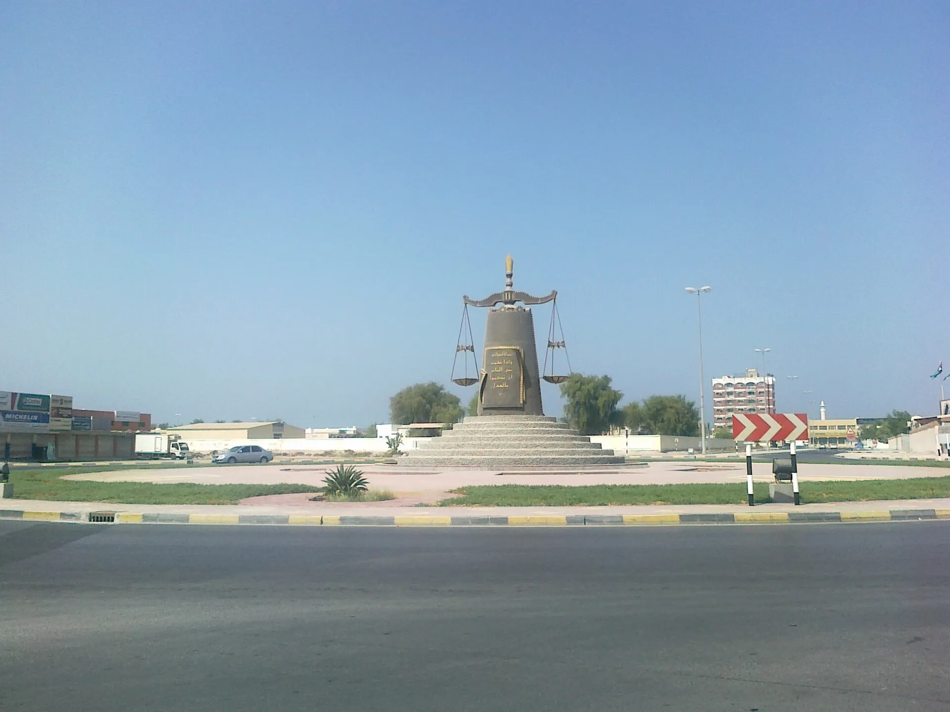 A scales of justice monument on a roundabout in Ras Al Khaimah, with low buildings and a clear sky behind