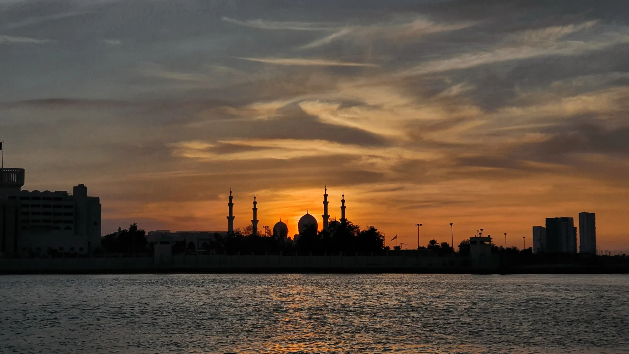 The minarets and domes of the Sheikh Zayed Grand Mosque in silhouette at sunset, across the water in Abu Dhabi