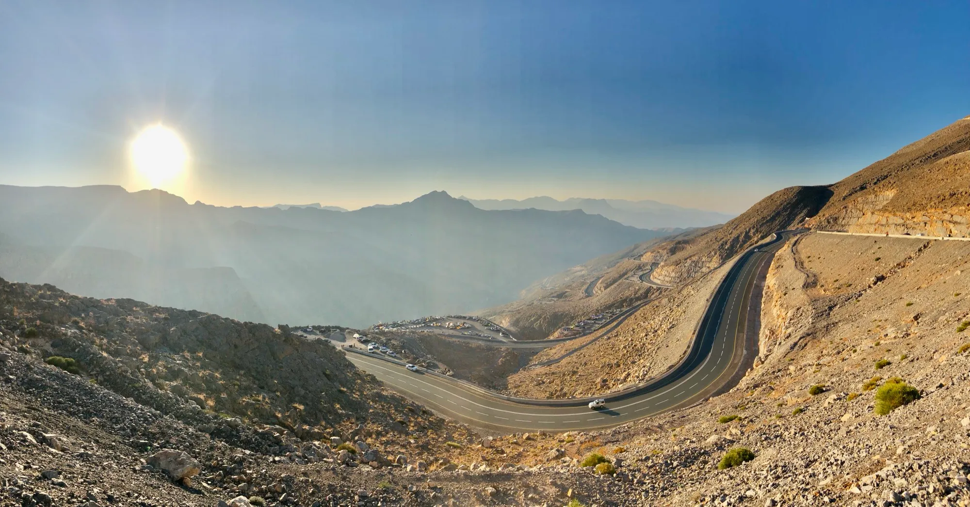 The Jebel Jais road curving down the mountainside past a viewpoint car park, with the low sun behind