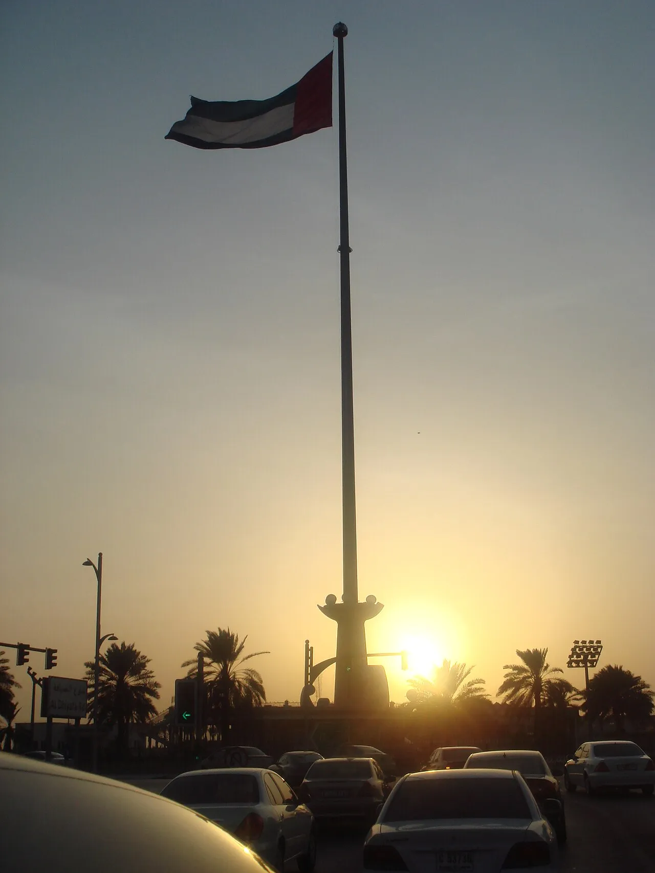 The UAE flag on a tall flagpole above evening traffic on a Jumeirah road in Dubai at sunset